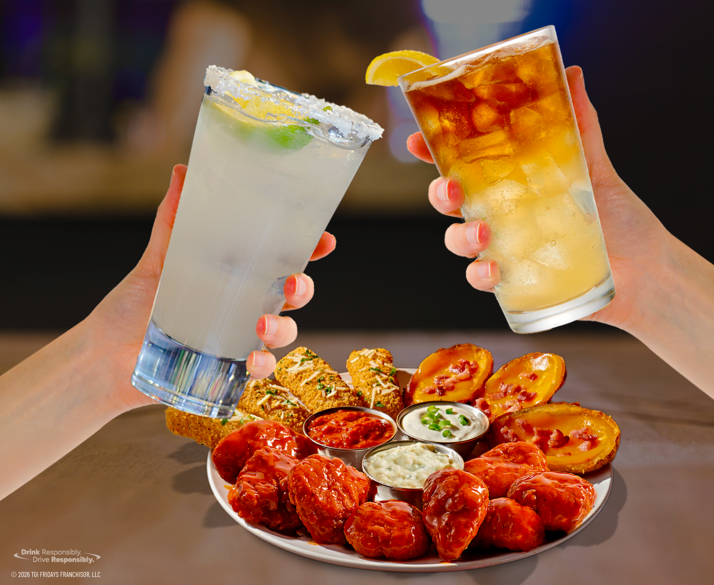 Two guests raising cocktails over a platter of boneless wings, mozzarella sticks, potato skins, and dipping sauces at TGI Fridays.